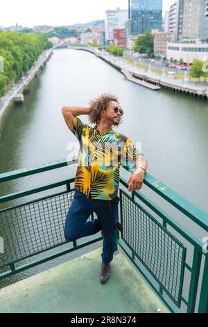 Passato di un uomo attraente con capelli afro in estate indossando una camicia di palma e la città sullo sfondo, godendo la vacanza Foto Stock