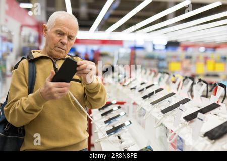 uomo anziano in grigio pensionato esaminano il contatore con gadget elettronici e tablet in showroom del negozio di merci digitali Foto Stock