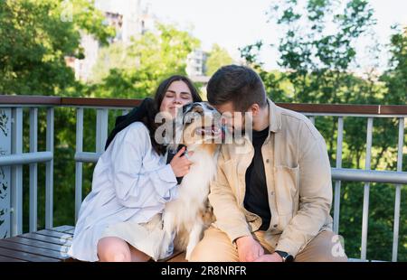 Giovane donna e uomo felici abbracciano il loro carino cane australiano in un parco. Cane da pastore australiano Blue merle nell'area del parco urbano accanto ai proprietari, che porta animali domestici per un Foto Stock