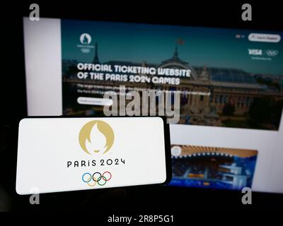 Persona che possiede il cellulare con il logo delle Olimpiadi estive di Parigi 2024 sullo schermo davanti alla pagina Web. Concentrarsi sul display del telefono. Foto Stock