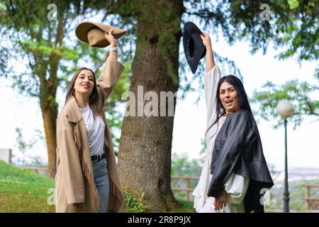 Due giovani amici femminili che si levano in piedi fuori in un paesaggio naturale, sorridendo e celebrando la loro amicizia alzando i loro cappelli nell'aria Foto Stock