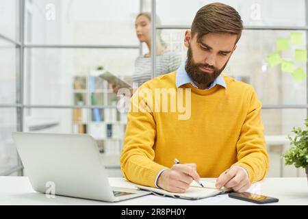 Serio occupato bel giovane uomo d'affari in giallo maglione scrivere idee in agenda mentre si lavora in ufficio moderno Foto Stock