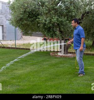 Immagine di un uomo che annaffia il suo giardino con un tubo. Cura e manutenzione del prato e riferimento ai rifiuti idrici e alla siccità. Foto Stock