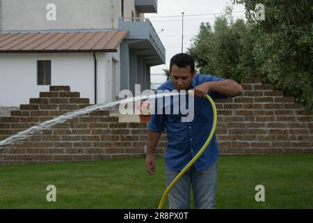 Immagine di un uomo nel suo giardino di casa che beve acqua da un tubo per prato. Riferimento ai rifiuti idrici. Foto Stock