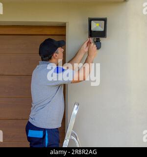 Immagine di un elettricista su una scala a forbice che utilizza i suoi strumenti per installare un riflettore su una casa. Illuminazione delle parti più buie della casa. Foto Stock