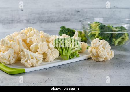 Broccoli florets in una ciotola di vetro e cavolfiori in primo piano su un tagliere sul tavolo da cucina Foto Stock