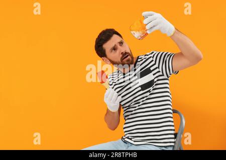 Designer emotivo con attrezzatura da pittura seduta su scala pieghevole vicino a parete arancione appena dipinta, spazio per testo Foto Stock