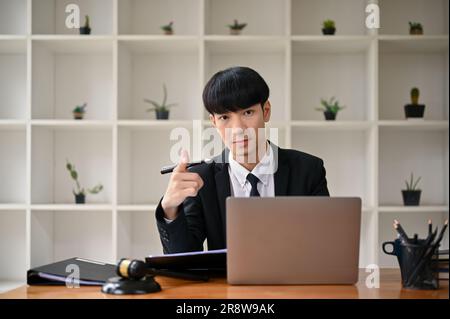 Un avvocato maschio asiatico, intelligente e professionale, con un vestito formale, punta il dito verso la telecamera mentre è seduto alla sua scrivania in ufficio. avvocato, avvocato, Foto Stock