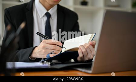 Immagine ritagliata di un avvocato maschio asiatico professionista in una causa formale che scrive qualcosa sul suo notebook mentre lavora alla sua causa alla sua scrivania in sala operatoria Foto Stock