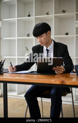 Un ritratto di un affascinante e professionale avvocato maschio asiatico millenario in una tuta formale sta scrivendo una lettera di avviso per il suo cliente alla sua scrivania in sala Foto Stock