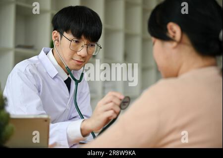 Un medico maschio asiatico millennial felice e professionale utilizza uno stetoscopio per esaminare il battito cardiaco di una paziente femminile in ospedale Foto Stock