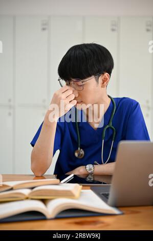 Un giovane studente di medicina asiatico maschio stressato e stanco in scrub sta lavorando alla sua ricerca medica a un tavolo nella sua stanza. Foto Stock