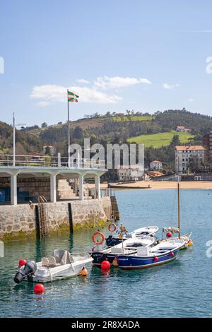 Lekeitio, Spagna - 30 marzo 2023: Parte del porto con barche ancorate, bandiera Euskadi, spiaggia, colline e foreste sullo sfondo Foto Stock