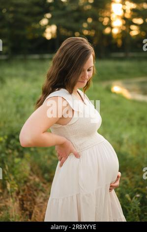 giovane donna incinta felice che guarda in basso e tiene la pancia nella foresta Foto Stock