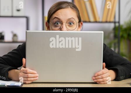 Donna d'affari sicura che si nasconde dietro un computer portatile, guarda la fotocamera, spiando i colleghi che lavorano, piangendo. Ragazza freelance professionista che guarda da dietro il computer con astuto sguardo occhi Foto Stock