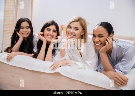 positività, festa nuziale, quattro donne, sposa felice e damigelle in abiti di seta sdraiati sul letto, guardando la macchina fotografica, diversità culturale, divertendosi a gattonare Foto Stock