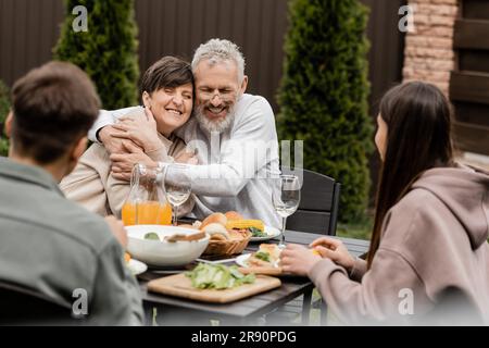 Allegra coppia di mezza età che si abbraccia vicino a bambini sfocati e cibo estivo durante la festa barbecue e la festa dei genitori nel cortile a giugno, giorno speciale per il barbecue Foto Stock