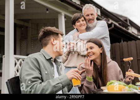 Sorridente ragazza adolescente che parla con il fratello mentre i genitori si abbracciano durante la festa al barbecue e la festa dei genitori nel cortile a giugno, giorno speciale per p Foto Stock