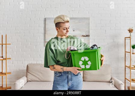 stile di vita eco-consapevole, donna giovane e tatuata con acconciatura alla moda che tiene una scatola verde di riciclaggio con abbigliamento, vita sostenibile e ambiente Foto Stock