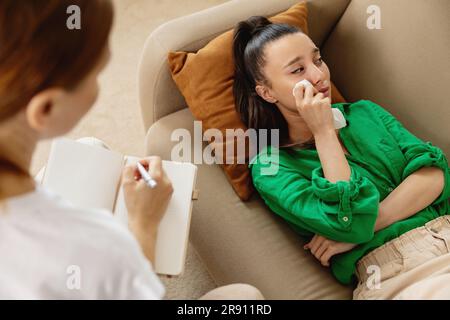 Vista posteriore del terapista che prende appunti mentre la paziente è sdraiata sul divano e parla dei suoi problemi. Foto Stock