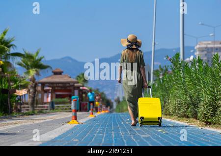 Una donna in abito verde cammina con una valigia gialla lungo il marciapiede. Foto Stock