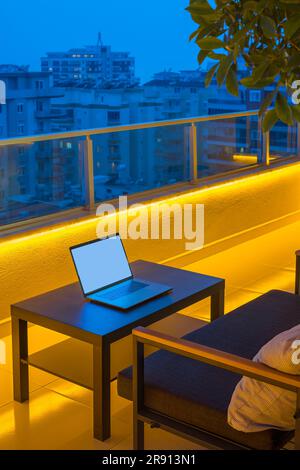 Un divano e un tavolino da caffè con un computer portatile si trovano su un ampio balcone di un alto edificio. Bella illuminazione gialla lungo il perimetro della Th Foto Stock