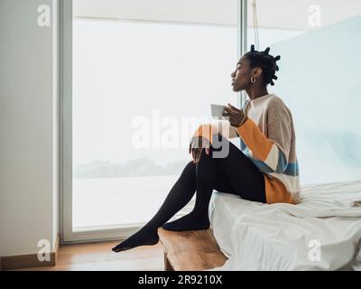 Giovane donna che beve un caffè seduto sul letto vicino alla finestra Foto Stock