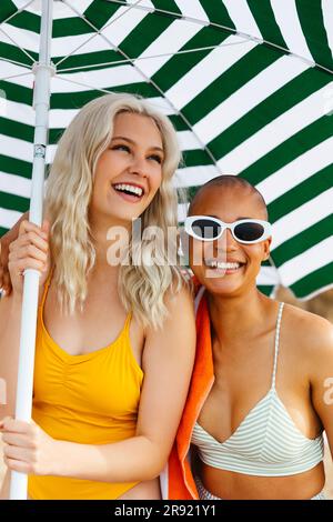 Amici sorridenti sotto l'ombrellone a strisce in spiaggia Foto Stock