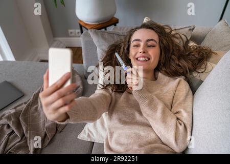 Giovane donna felice che mostra il kit del test di gravidanza in videochiamata a casa Foto Stock