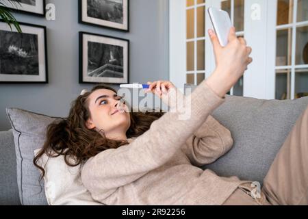 Donna sorpresa che mostra il kit del test di gravidanza in videochiamata a casa Foto Stock