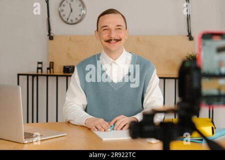 Uomo sorridente seduto in classe che guarda la fotocamera che registra una lezione online Foto Stock