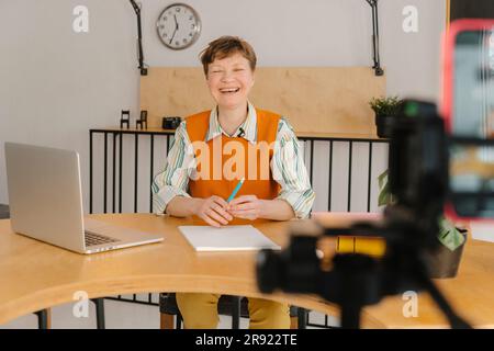 Donna sorridente seduta in classe che guarda una videocamera che registra una lezione online Foto Stock