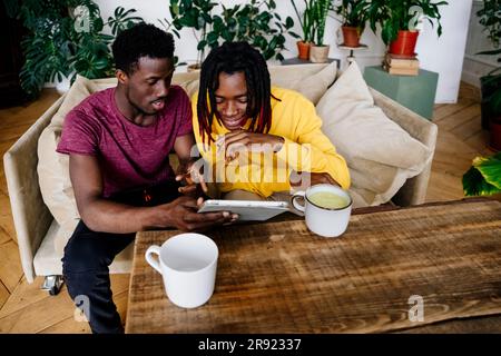 Uomo sorridente che condivide un tablet PC con un amico seduto a casa Foto Stock