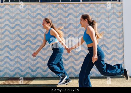 Sorelle che si tengono per mano e corrono davanti al muro Foto Stock