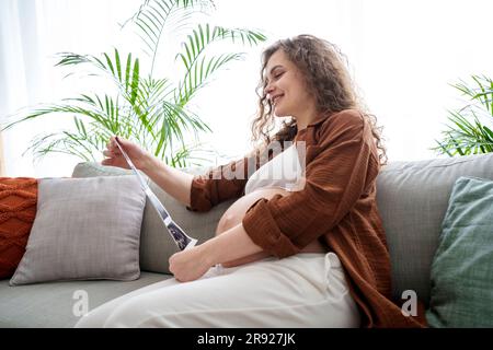 Donna incinta sorridente che guarda le foto ecografiche seduto sul divano Foto Stock