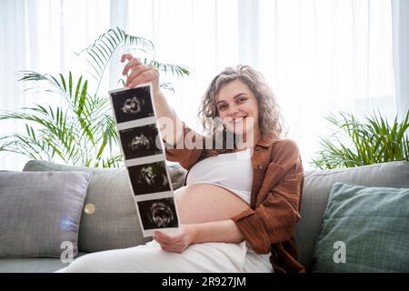 Donna incinta sorridente che mostra ecografie seduto sul divano a casa Foto Stock