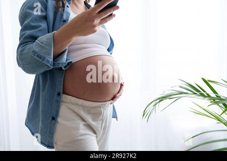 Giovane donna incinta che tocca la pancia e usa lo smartphone a casa Foto Stock