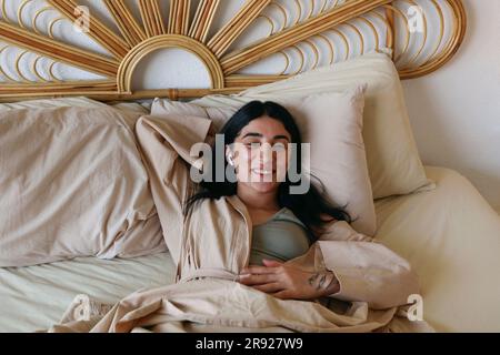 Giovane donna sorridente che si rilassa a letto e ascolta la musica a casa Foto Stock