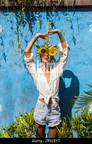 Donna sorridente che nasconde il volto con i girasoli davanti al muro Foto Stock