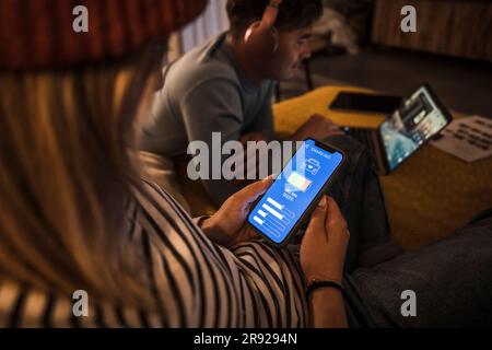 Giovane donna che utilizza l'app di ricarica per auto elettriche sullo smartphone e uomo che utilizza un computer portatile in background Foto Stock