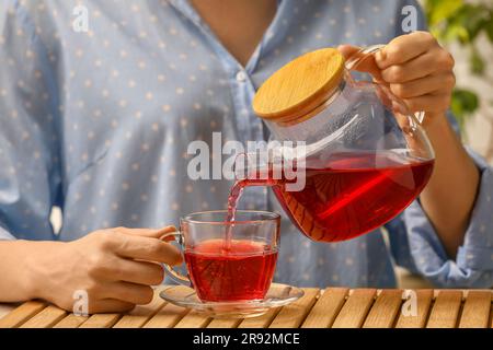 Donna che versa delizioso tè all'ibisco in una tazza al tavolo di legno, primo piano Foto Stock