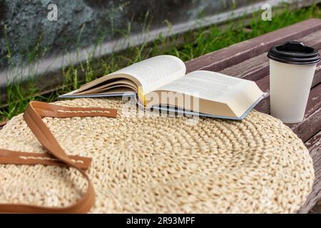 Aprire il libro con la tazza di caffè sul sacchetto di paglia. Tempo libero nel parco. Libro aperto e caffè da portare sulla panchina. Rilassati in estate. Stile di vita accogliente. Leggendo fuori. Foto Stock