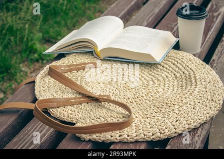Aprire il libro con la tazza di caffè sul sacchetto di paglia. Tempo libero nel parco. Libro aperto e caffè da portare sulla panchina. Rilassati in estate. Stile di vita accogliente. Leggendo fuori. Foto Stock