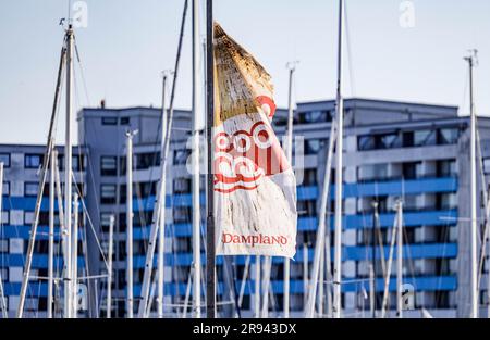 PRODUZIONE - 08 giugno 2023, Schleswig-Holstein, umido: Una bandiera con l'iscrizione Dampland wehr davanti al porto di fronte agli appartamenti per le vacanze in umido su un palo. Negli anni '1960 e '1970 furono create le strutture di base per il turismo moderno nello Schleswig-Holstein. Dove oggi le famiglie giocano a minigolf, i marinai saltellano nel porto e le coppie godono di un trattamento benessere, è stato un bel 50 anni fa: Praticamente nulla. Ma le cose cambiarono presto. Nel mezzo di campi, prati e Mar Baltico, un centro vacanze con il nome futuristico "Dump 2000" è stato costruito dal 1969 al 1973 nel comune Foto Stock