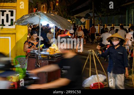 Una donna che vende spuntini con la sua bancarella all'angolo di una strada tra una miriade di locali e turisti di notte nella città vecchia di Hoi An, in Vietnam. Foto Stock