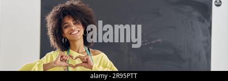 Il giovane tutor afro-americano in maglione giallo brillante mostra il cuore con le dita alla classe di cucito Foto Stock