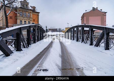 Klodzko, Polonia - gennaio 2023: Piccolo ponte con parti di costruzione in metallo nero sopra un piccolo fiume pieno di neve in inverno Foto Stock