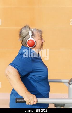 Vista del profilo di una donna anziana obesa attiva in abbigliamento sportivo che si allena su una macchina da palestra in una palestra del parco ascoltando musica con le cuffie Foto Stock