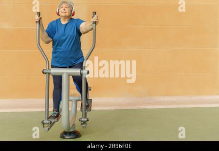 Donna anziana obesa attiva in abbigliamento sportivo che si allena su una macchina da parcheggio mentre ascolta la musica sulle cuffie Foto Stock