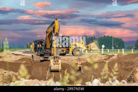 Escavatore, pala e geometra che lavorano su un cantiere stradale al tramonto estivo, con erba in primo piano Foto Stock